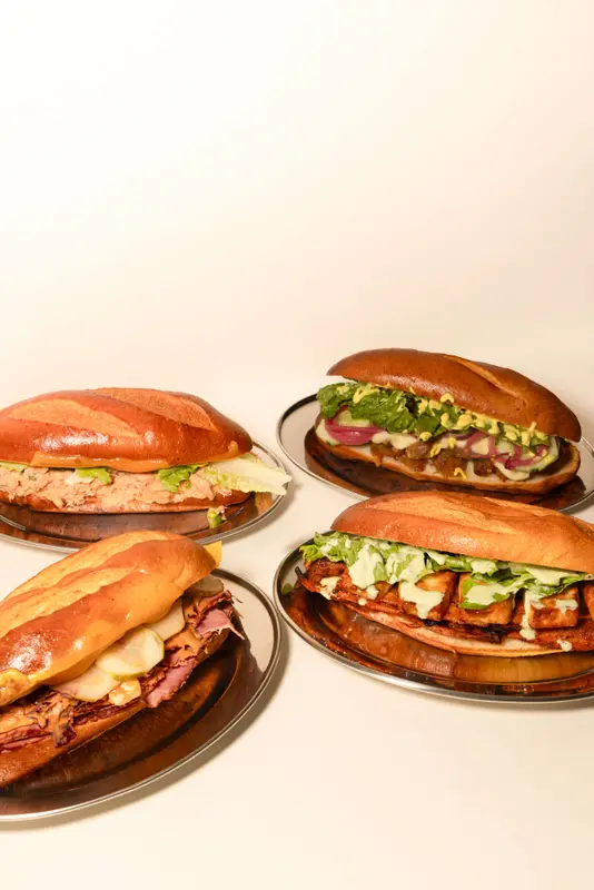Four gourmet submarine sandwiches on silver plates featuring tuna, BBQ tofu, pastrami, and a veggie option with fresh toppings.