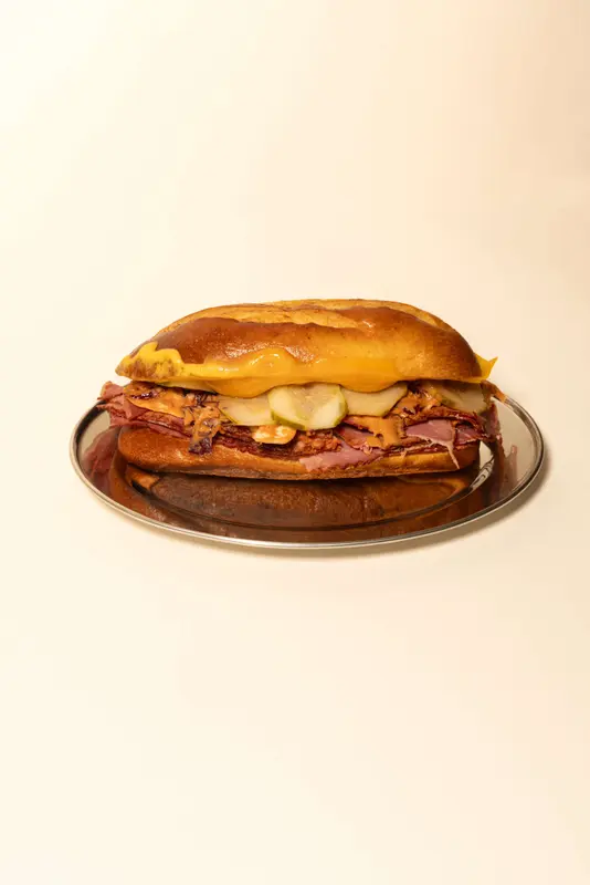Toasted hoagie sandwich with pastrami, melted cheddar, pickles, and mustard on a silver plate