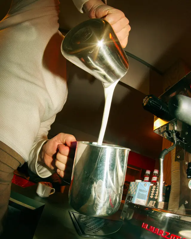 Barista pouring steamed milk into pitcher on La Marzocco espresso machine at specialty coffee bar