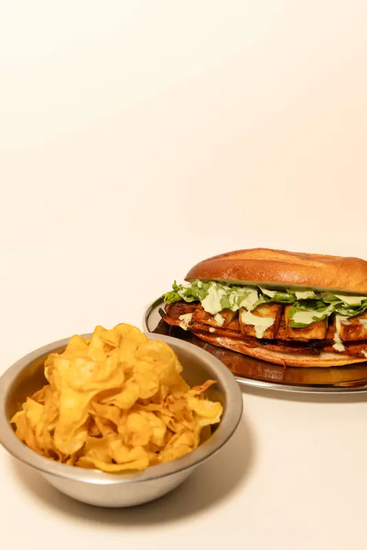 Grilled tofu sandwich with lettuce and aioli on brioche bun, served with crispy potato chips in a steel bowl.