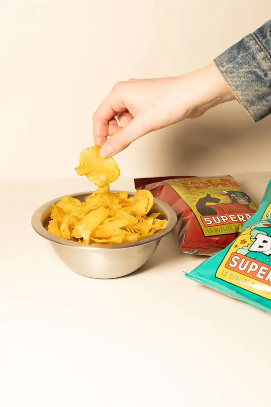 Hand grabbing a salt and pepper potato chip from a stainless steel bowl beside Superb brand chip bags.
