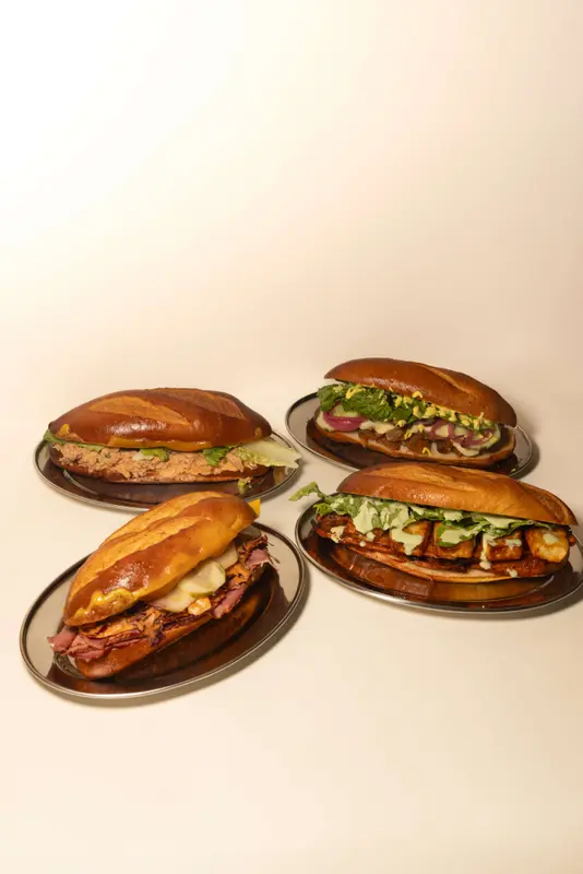 Four gourmet sandwiches on pretzel rolls served on silver plates, filled with meats, melted cheese, pickles, and fresh greens.
